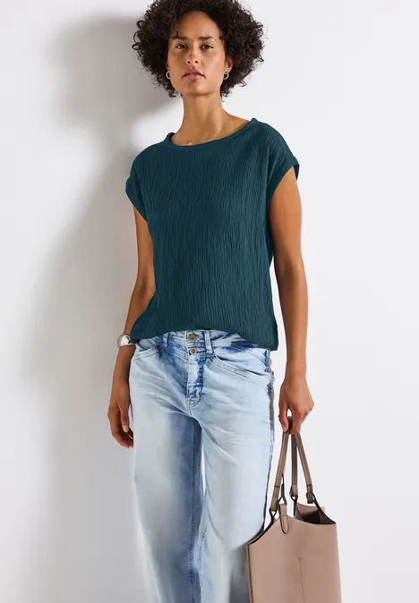 Frau mit lockigem Haar, grünem Shirt, heller Jeans und beigefarbener Tasche, steht vor weißer Wand.